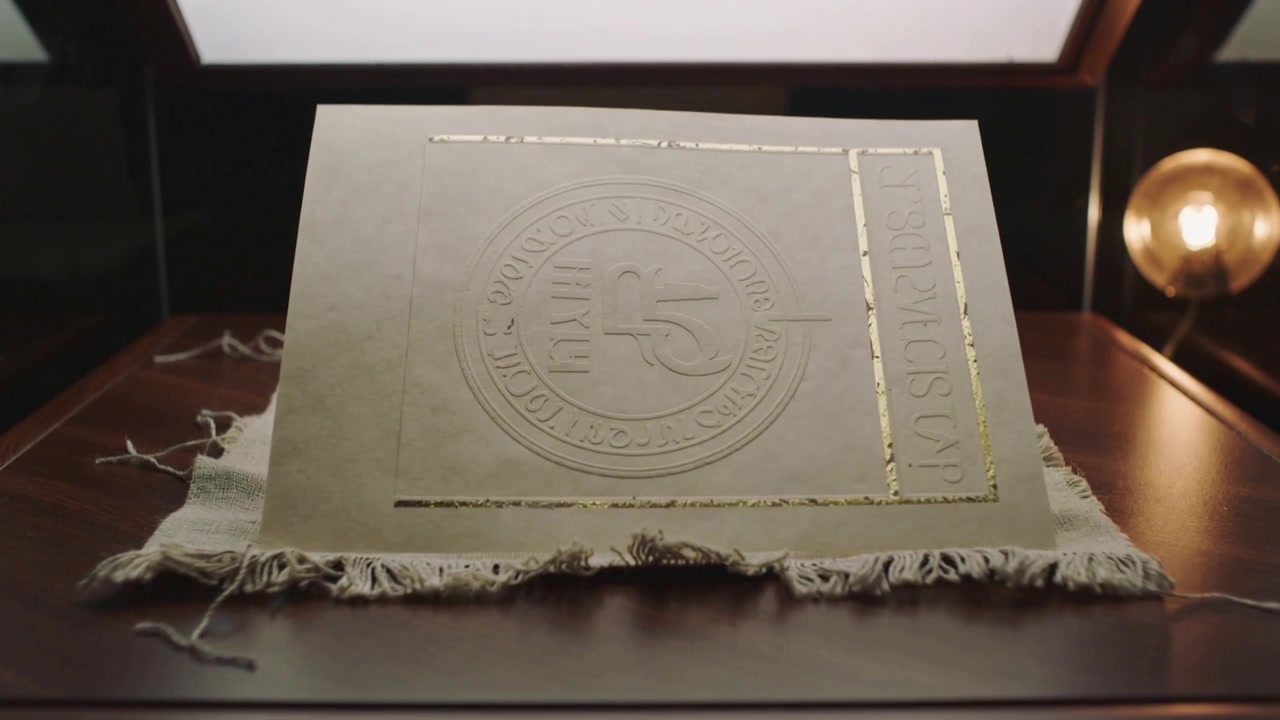 Carte gaufrée inclinée, le bord se soulevant et tournant sur un tapis effiloché dans une vitrine en verre, révélant un sceau doré.