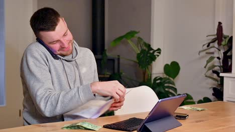 Joyful-white-young-man-reviews-bill-on-paper-and-talks-on-phone,-seated-at-a-table,-nonverbally-expressing-his-satisfaction-with-a-contented-smile