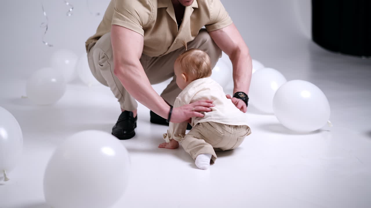 Man picking a little baby sitting on the floor. Father tosses his lovely child up and kisses him lovingly. Studio footage with balloons around.