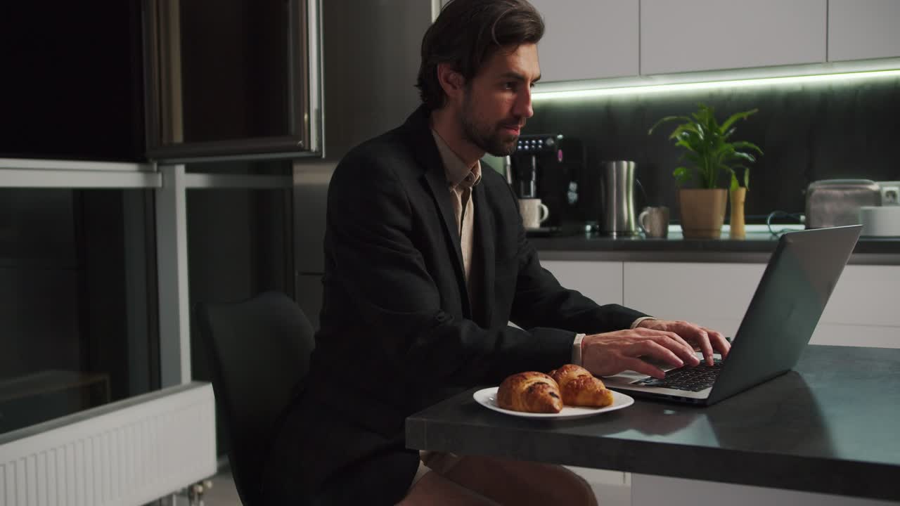 A confident brunette man in a black jacket and beige shorts works at a gray laptop and communicates via video in a modern kitchen in the evening. Confident brunette man with stubble communicates via video call while sitting in only beige shorts in the kitchen in the evening