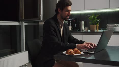 a confident brunette man in a black jacket and beige shorts works at a gray laptop and communicates via video in a modern kitchen in the evening. confident brunette man with stubble communicates via video call while sitting in only beige shorts in the kitchen in the evening