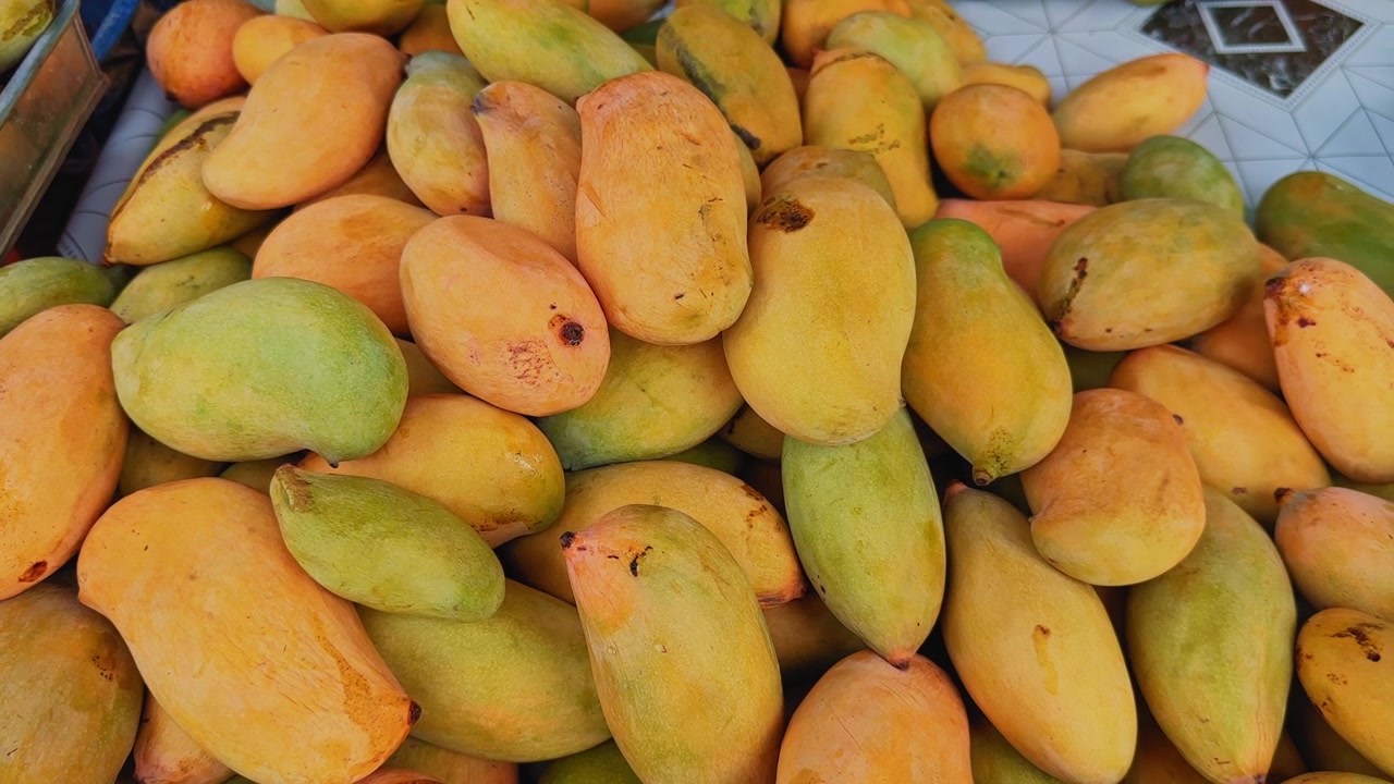 Płynne ujęcie z jazdy przedstawiające świeże, dojrzałe mango ułożone w stos na lokalnym targu owocowym, podkreślające naturalne kolory, sezonowe zbiory i tropikalne produkty