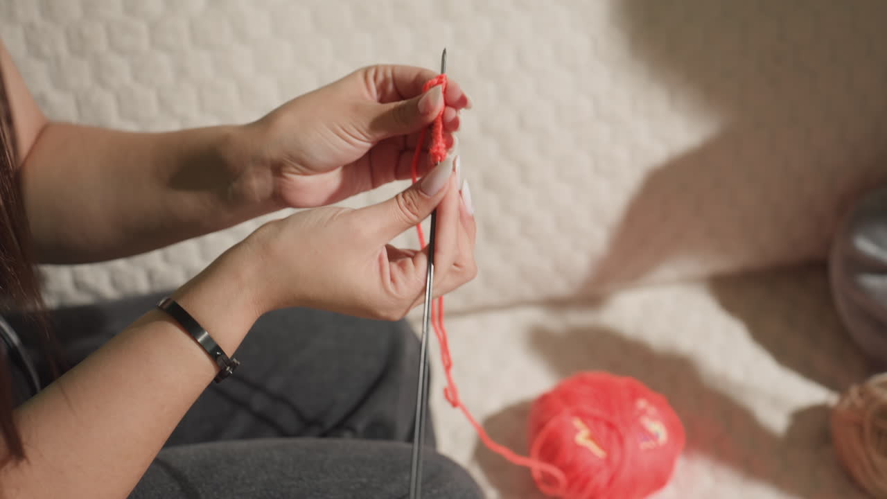 백인 여성이 부드러운 침대에서 차분하게 뜨개질로 테라피를 하고 있어요. 손을 가까이 대고 천천히 움직이며 빨간색 스티치를 만들고, 아늑한 쿠션과 따뜻한 색조가 스트레스 해소를 위한 치료 공예 분위기를 조성합니다.