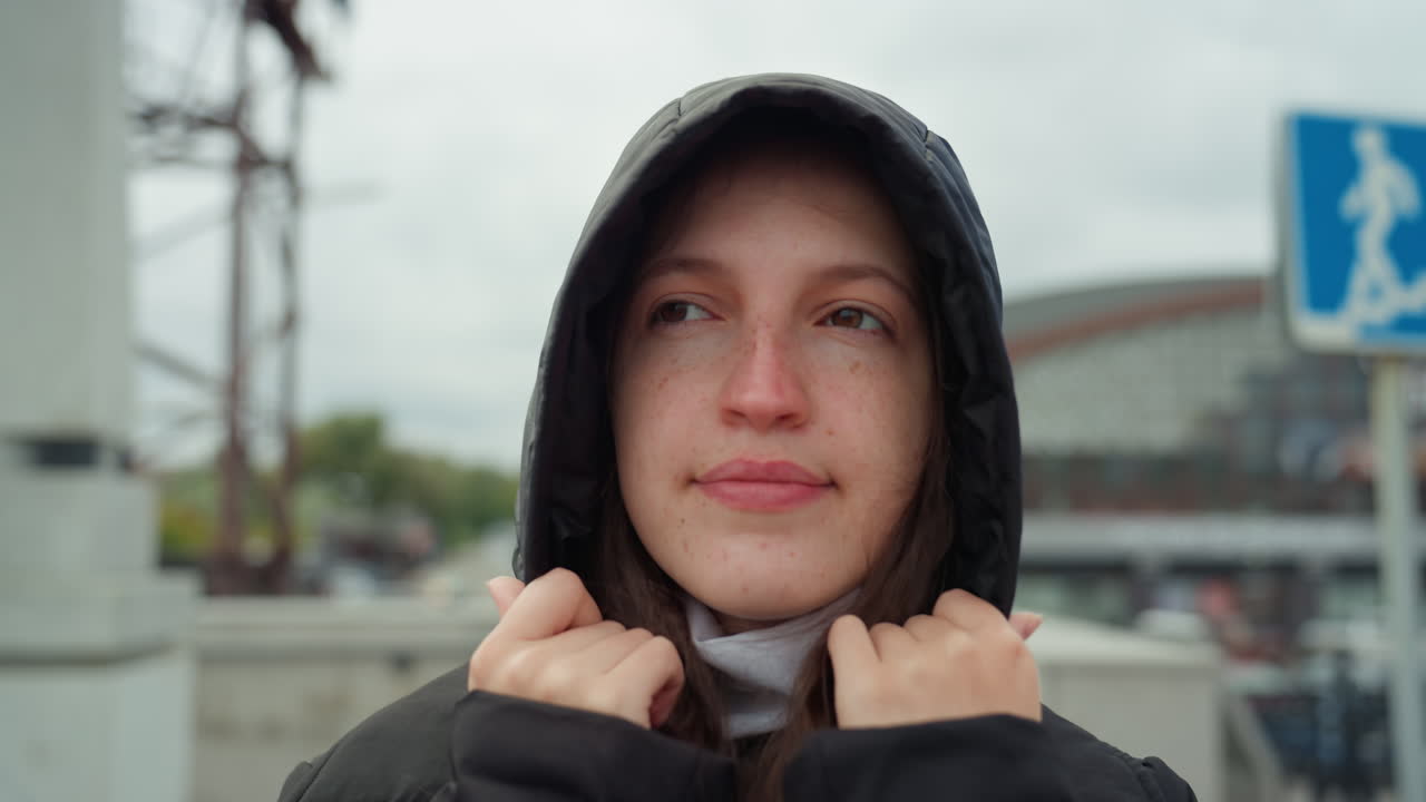 Nuori valkoihoinen nainen säätämässä huppua ulkona, pilvinen taivas, kävelysilta lähellä rautatieasemaa, musta takki, kädet pitelevät hupun reunoista, hillitty hymy, hidas kävelyvauhti, avoin muotokuvatyyli, kaupunkimainen