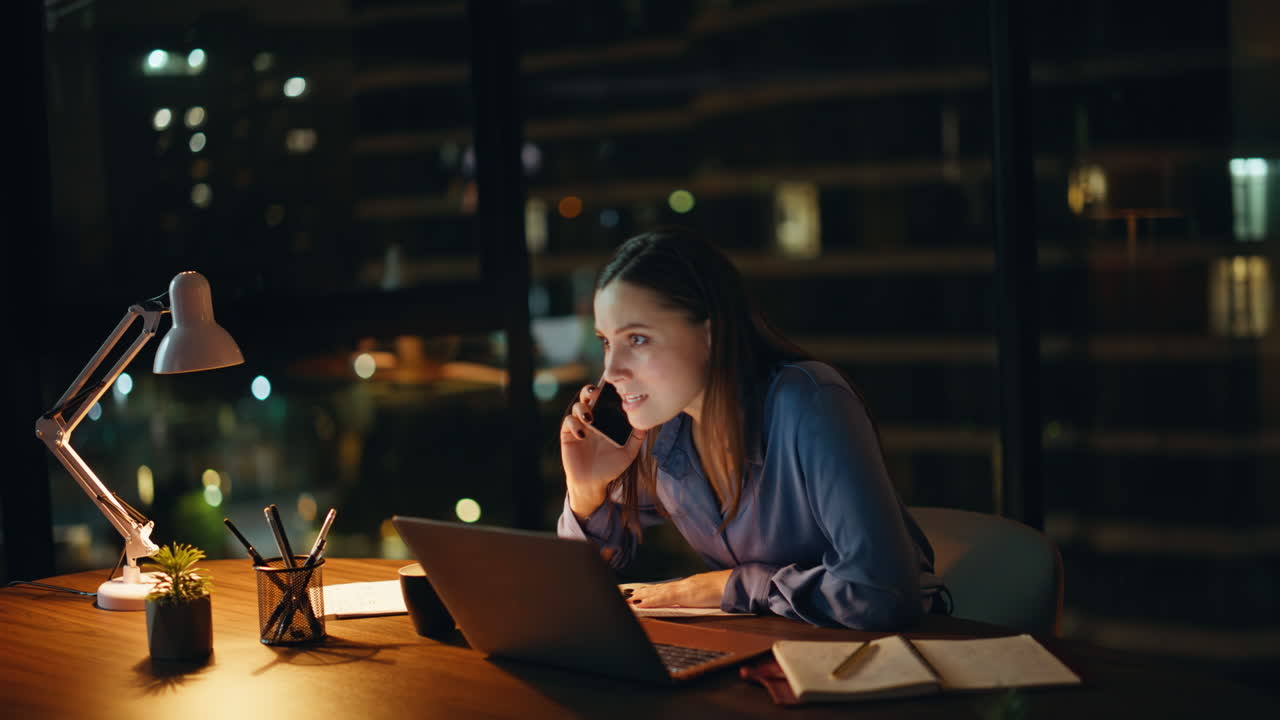 Overtime manager talking cellphone looking laptop at night office. Worried woman