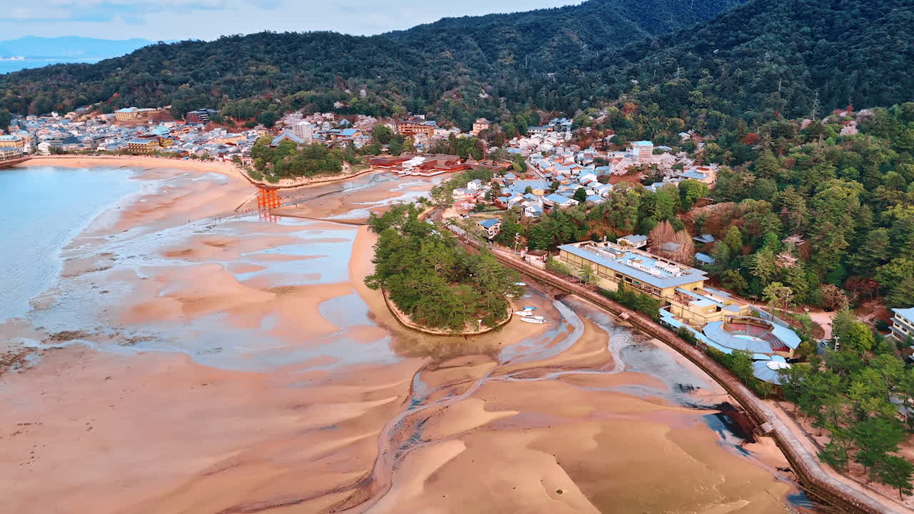 Beautiful scenery of the sandy beach at the foot of the wooded green mountains. Drone footage approaching Itsukushima Shrine at low tide season. Stunning view of the Miyajima island, Japan.