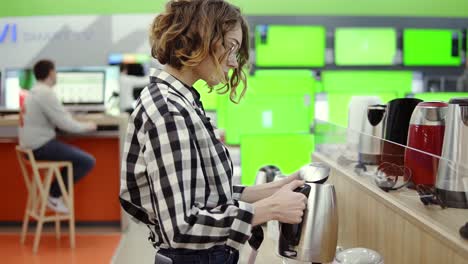 Side-View-Of-A-Young-Cheerful-Positive-Attractive-Woman-In-Plaid-Shirt-Choosing-Electronic-Kettle-In-Household-Appliances-Store,-Standing-In-A-Row,-Holding-And-Examines-Silver-Kettle