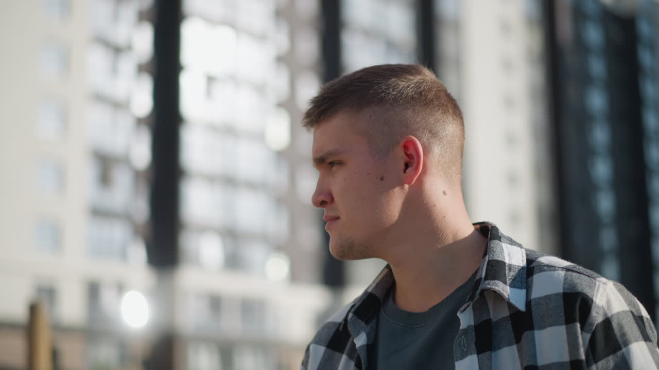 Side profile of thoughtful student in checkered shirt meditating outdoors during sunny day with soft sunlight reflecting on face and blurred view of tall modern high-rise building in urban background