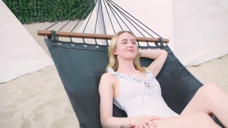 sexy caucasian blonde young girl woman lie down in a hammock wearing one-piece swimsuit in sandy tropical beach, relaxing enjoying her holiday in modern luxury resort spa