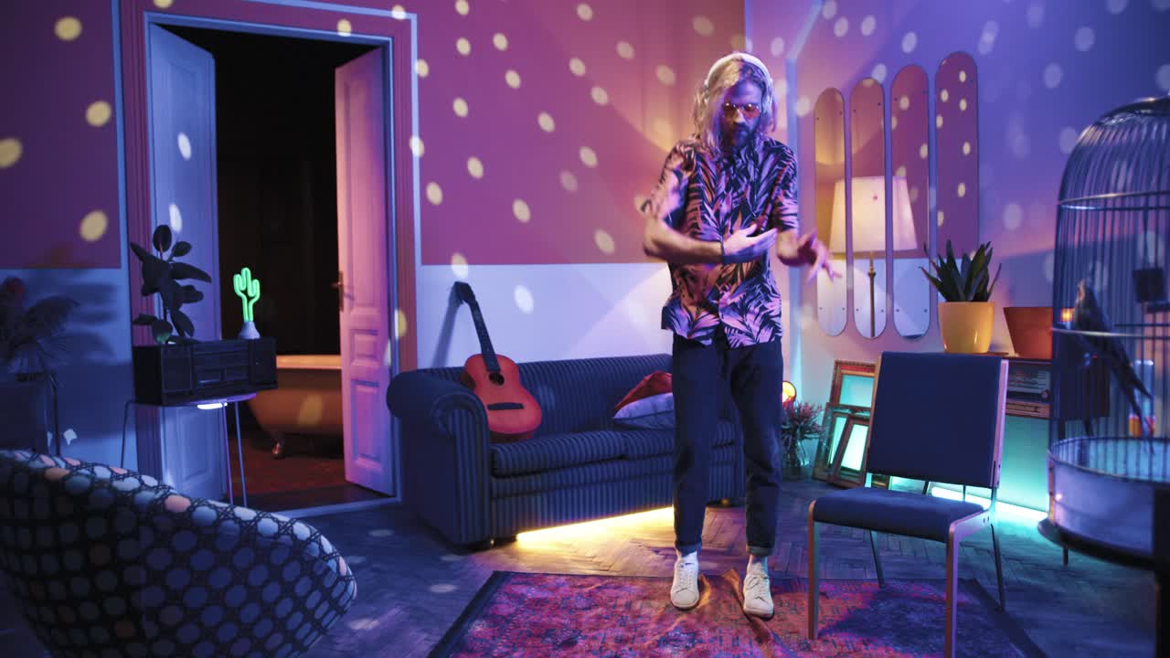 헤드폰을 끼고 긴 머리를 하고 집에 있는 복고풍 파티에서 정력적으로 춤을 추는 안경을 쓴 젊은 세련된 백인 남자