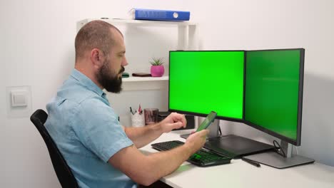 hombre sentado en casa y enviando mensajes de texto en el teléfono inteligente contra dos pantallas o monitores verdes. dedos escribiendo en la pantalla táctil