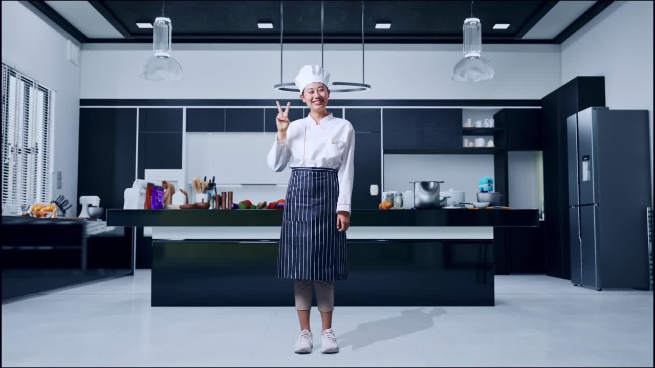 아시아 여성 셰프의 몸이 가득 차 있고 미소 짓고 가정 부에서 카메라에 평화의 몸짓을 보여줍니다.