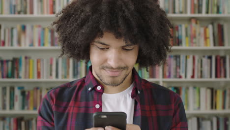 Porträt-Junger-Mann,-Student,-Der-Smartphone-Bücherregal-Bibliotheksuniversität-Nutzt