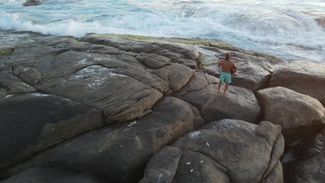 toma aérea en órbita de un joven tocando una canción emocional en la guitarra durante la hermosa puesta de sol en la playa con olas rompiendo en australia