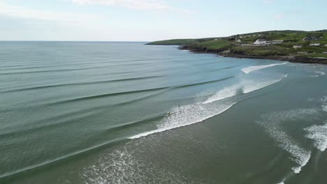 Drone-Volando-Hacia-Atrás-Sobre-Olas-Y-Surfistas,-Revelando-Más-Del-Paisaje-En-West-Cork,-Irlanda.