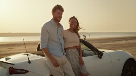 a blonde woman in a white top together with a bearded man in a blue shirt leaning on a white convertible car look into each other's eyes and enjoy the view on the river bank