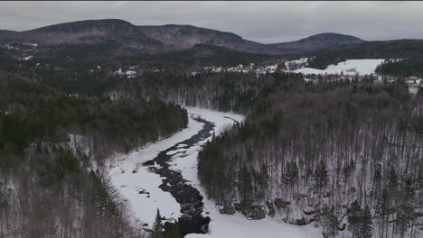 Aerial-scenic-drone-pan-up-and-forward-Canadian-wilderness-mid-winter-near-north-Quebec-Stoneham-Ski-Resort-of-frozen-over-Sautaurski-River-with-cozy-cabin-and-chalets