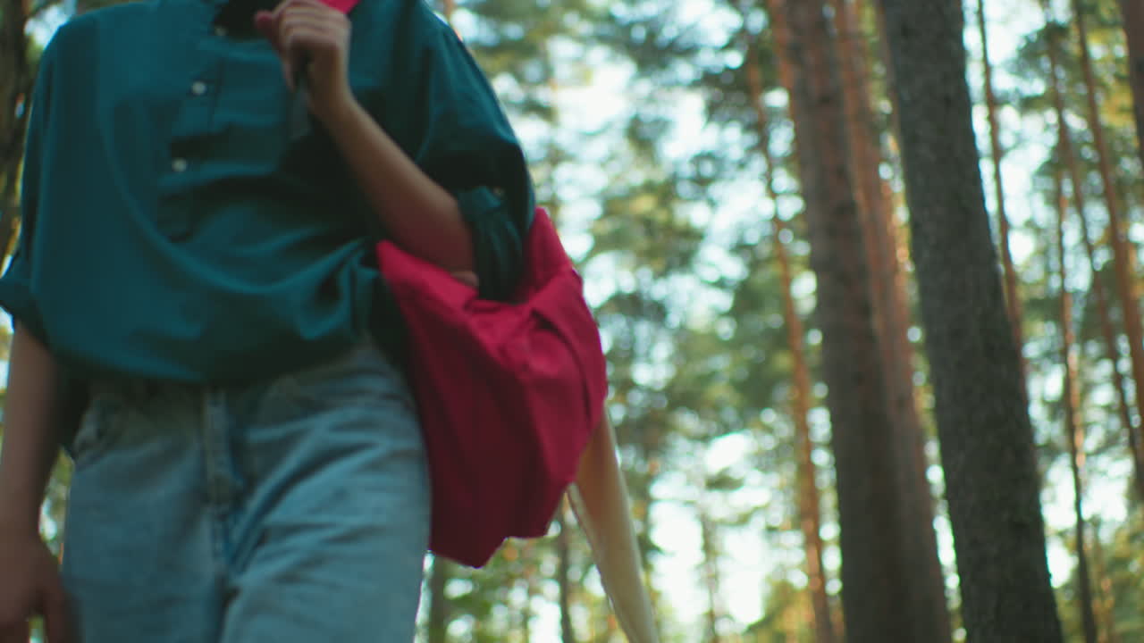 높은 숲을 가로질러 하이킹하는 여성의 낮은 각도 뷰, 어깨에 빨간 배을 들고, 자연 주변을 둘러보며, 빛이 나무를 통해 필터링됩니다.