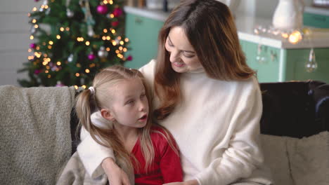 mother and daughter sitting on the sofa covered by a blanket while talking in living room with christmas decorations 2