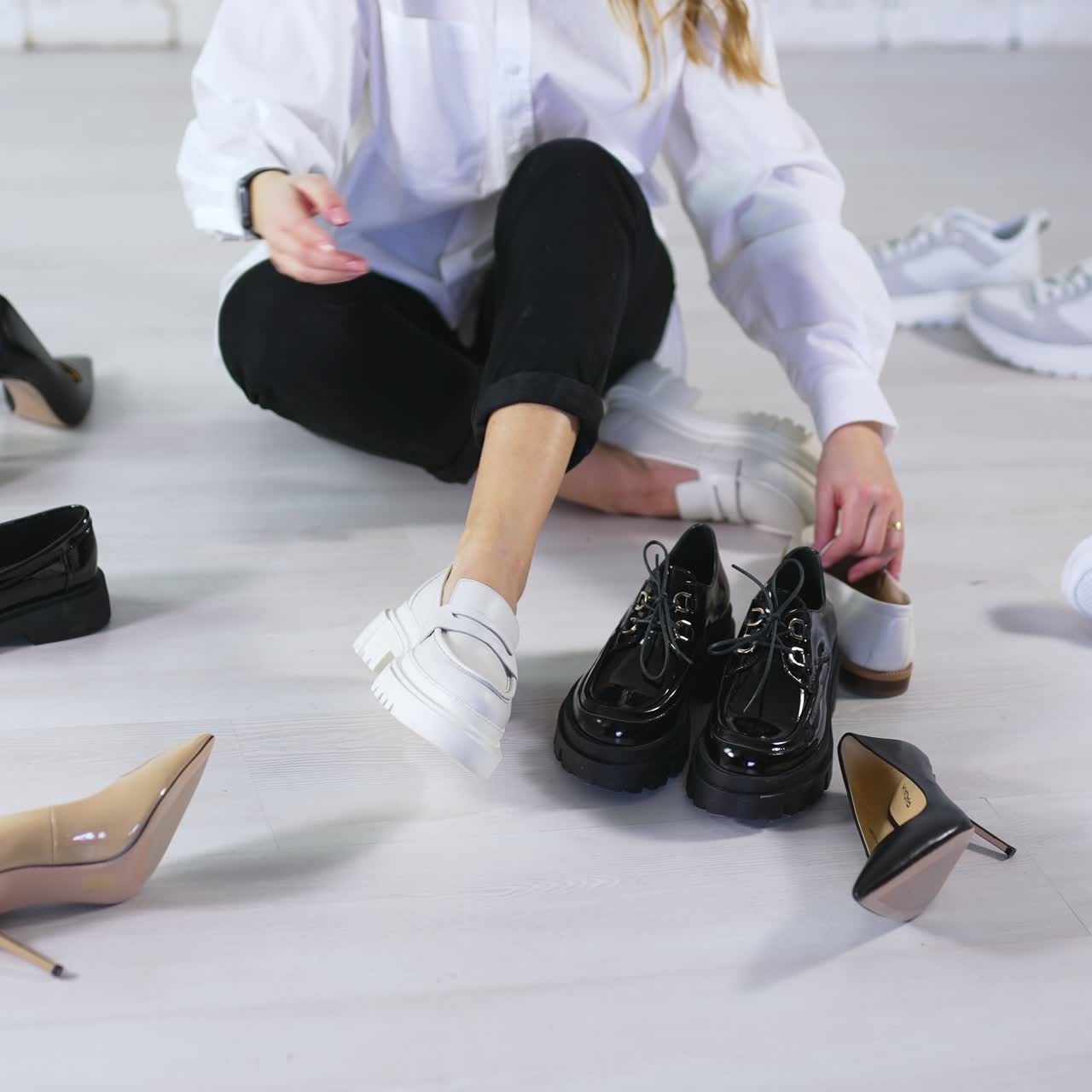 Looking through diverse pair of modern fashionable footwear. Lady sits on the floor in white shoes and takes other footgear to see