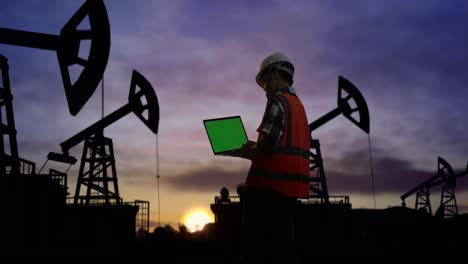 vista trasera de un ingeniero masculino asiático con casco de seguridad trabajando en una computadora portátil de pantalla verde y mirando a su alrededor mientras está de pie frente a las bombas de aceite, durante la puesta o el amanecer