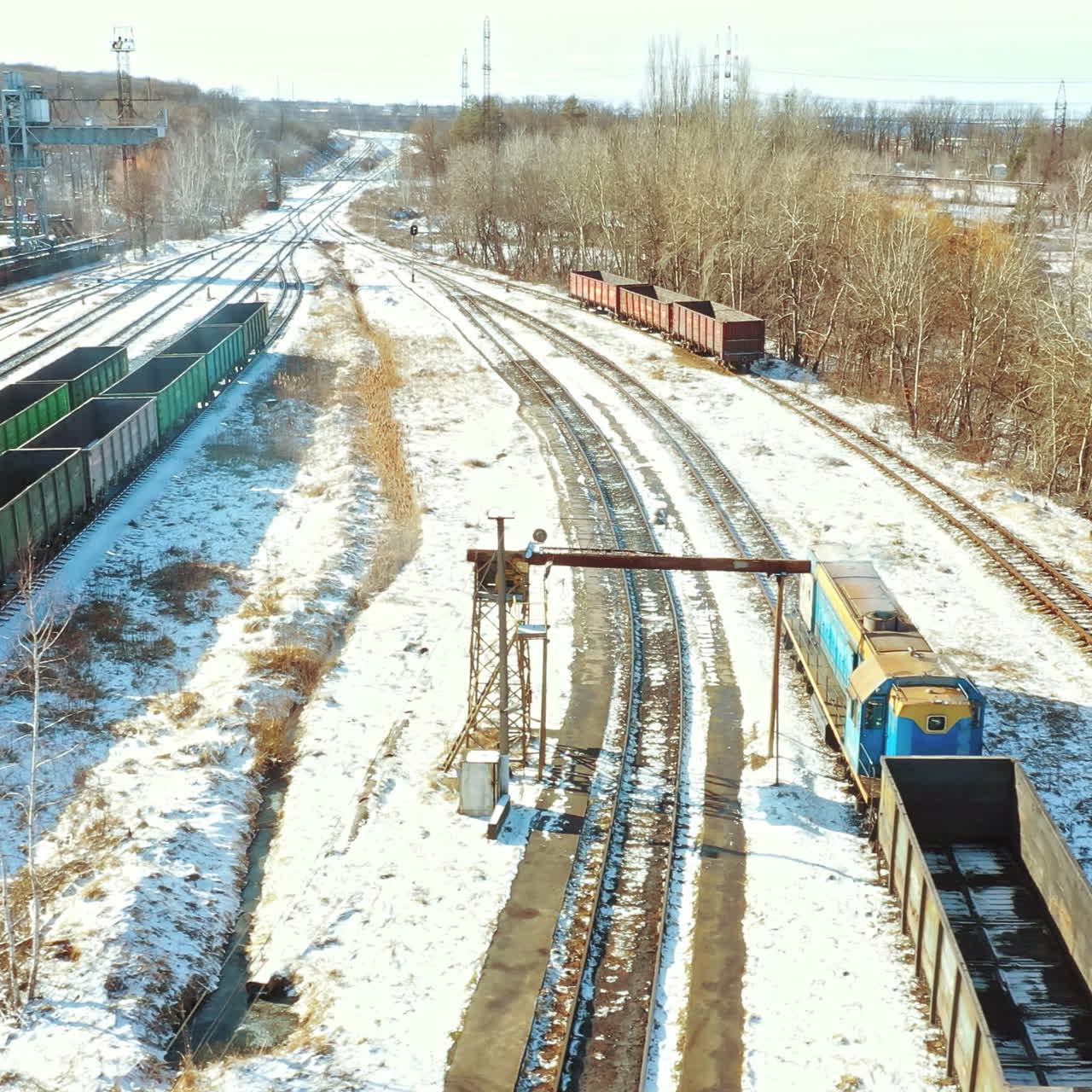 A freight train pulls a trailer with containers behind on the railroad in the background of the square of rail station with a multitude rows of container on the outskirts of the city in the winter. Aerial view.