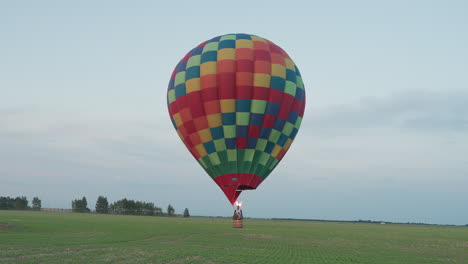 rear view passenger riding colorful hot air balloon gliding low across vast green farmland at dusk fueled by fiery burner against pale sky with distant tree line