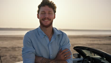 portrait: a bearded man with curly hair wearing a blue shirt is crossing his arms, smiling and standing next to a white convertible against the yellow sky on the beach