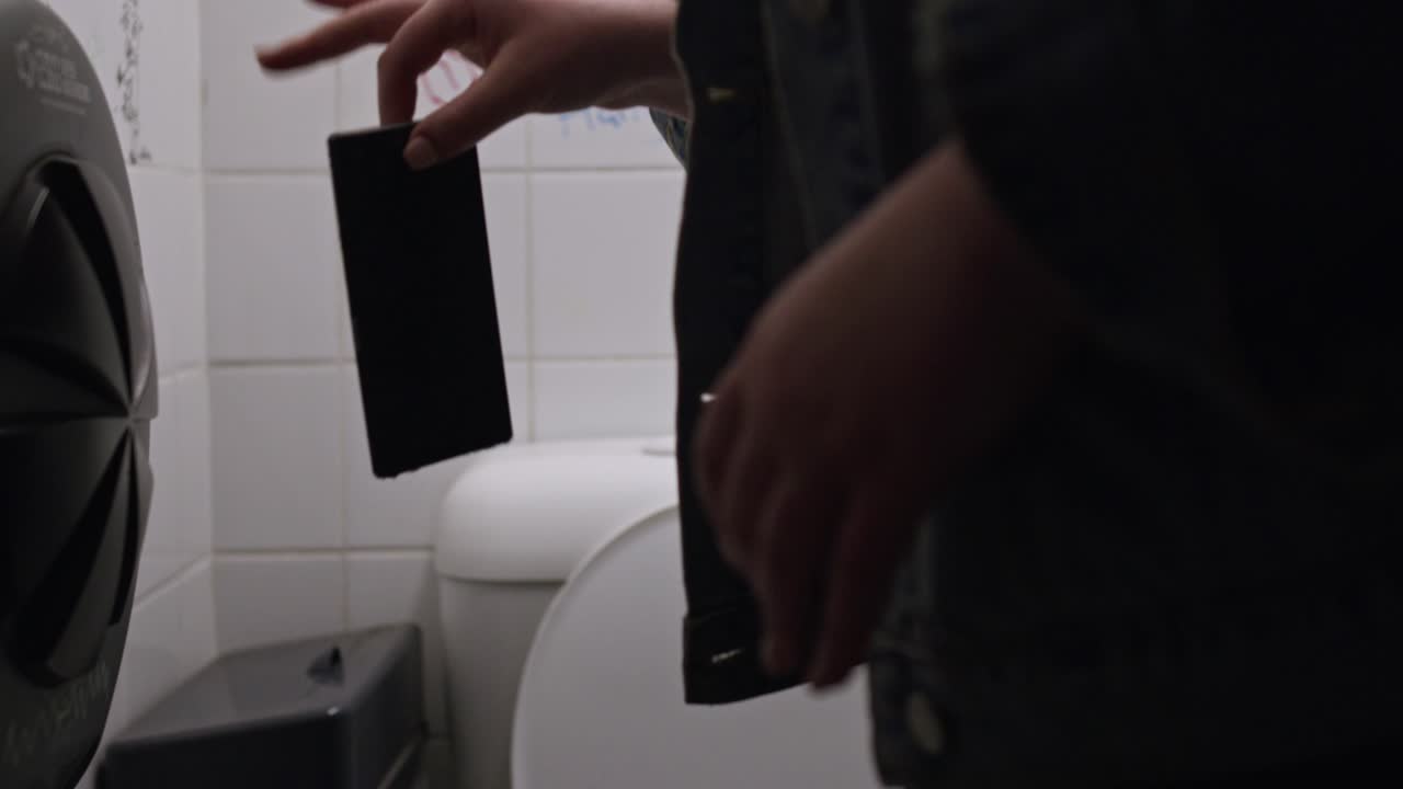 A young woman accidentally drops her phone into a toilet in a dimly lit bathroom, reacting with shock and frustration as it slips from her hand — close-up