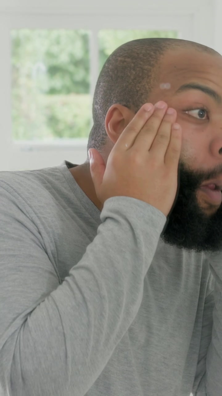 Vertical video: Checking mirror, man in gray shirt applying to beard with hands at vanity, readying