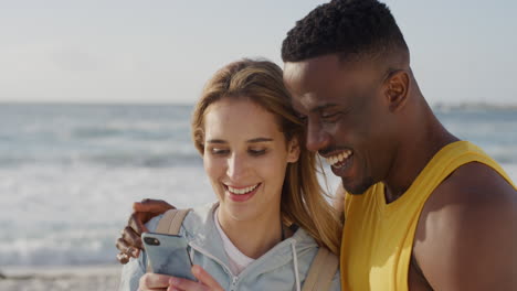 Porträt-Eines-Jungen-Multiethnischen-Paares,-Das-Mit-Dem-Smartphone-Am-Strand-SMS-Nachrichten-Liest-Und-Lächelt-Und-Gemeinsam-Einen-Romantischen-Urlaub-An-Der-Warmen-Sommerküste-Genießt