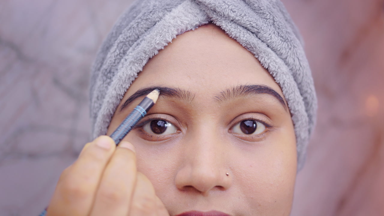 Close up shot of an Indian woman applying brow pencil on eyebrows during daily makeup routine indoor.
