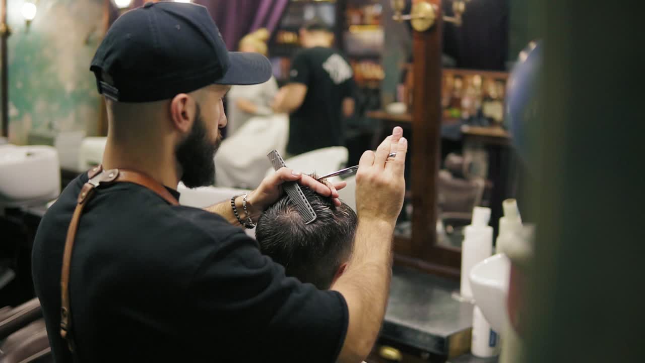 Back view of the stylish bearded barber in a black cap wearing leather apron performing a haircut with scissors holding a hair
