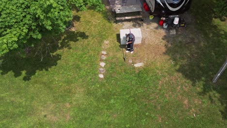 Blick-Von-Oben-Auf-Einen-Männlichen-Camper,-Der-Auf-Dem-Campingplatz-Gras-Mit-Einem-Freischneider-Trimmt