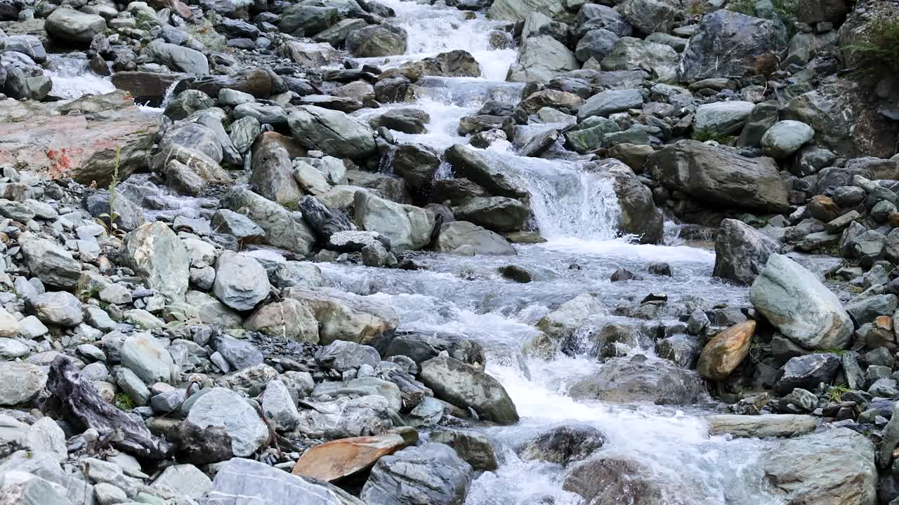 Clear mountain stream flows rapidly over large rocks and boulders in a natural outdoor setting, captured with steady camera and soft daylight
