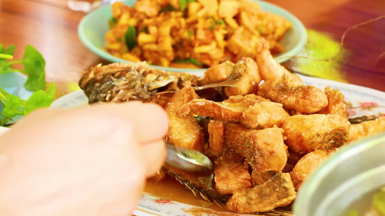 A hand serves deep-fried fish with sauce, surrounded by vibrant Thai dishes, in a warmly lit setting
