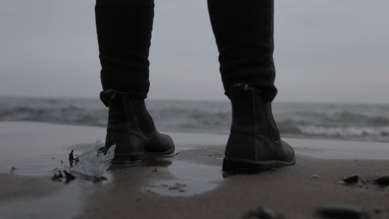 A Person Wearing A Black Leather Boots Standing In The Shoreline - Close Up Shot