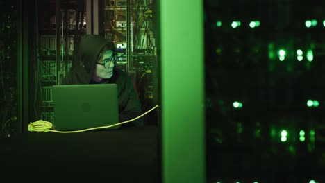 asian male hacker in hoodie using laptop by computer servers