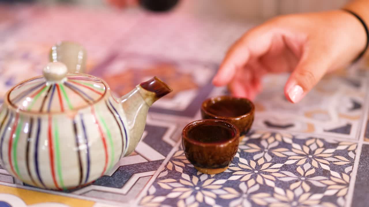 A hand pours tea from a striped teapot into small cups on a patterned tablecloth, creating a calming atmosphere