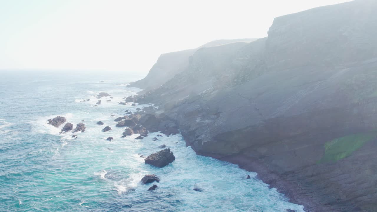 스페인의 playa de tagle의 산악 해안선의 능선과 정상에 있는 바다 파도, 안개가 가득한 하늘에서 태양이  ⁇ 아오르는 영화적인 공중 촬영