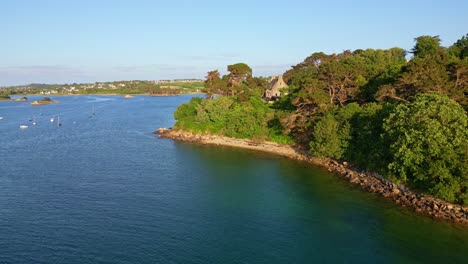 Backward drone movement from the wooded bay part jutting into calm blue seawater under evening sunlight at Port Blanc, Côtes-d'Armor, Brittany, France.