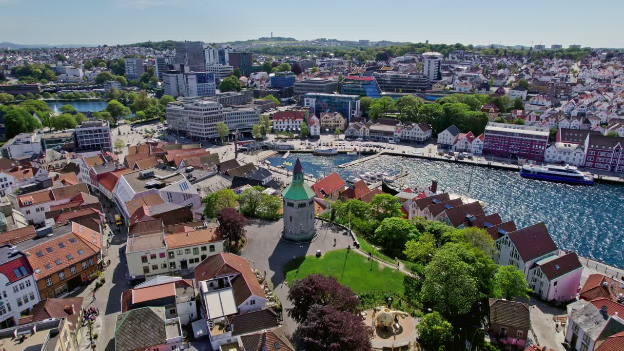 Drone circles Valberg Tower, showcasing its historic architecture with Vågen harbor and Stavanger cityscape in the sunny background.