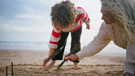 Mamá-Niño-Dibujando-Arena-De-Playa-En-El-Cielo-Nublado.-Familia-Creativa-Divirtiéndose-Juntos