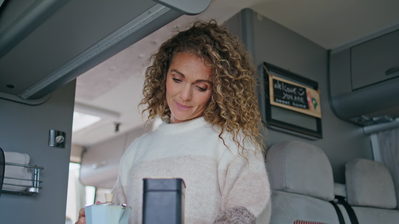 캠핑 밴에서 커피를 준비하는 곱슬한 여인. 관광객이 아침 식사를 하고 있다.