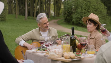 middle aged man plating a guitar sitting at table with his friends during an outdoor party in the park 1