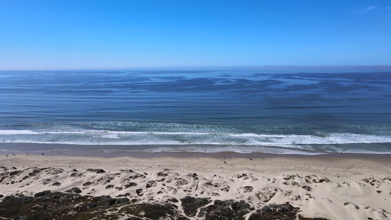 experimente la tranquila belleza de la costa con esta impresionante vista de las olas azules del océano, el cielo despejado y las dunas de la playa de arena en primer plano