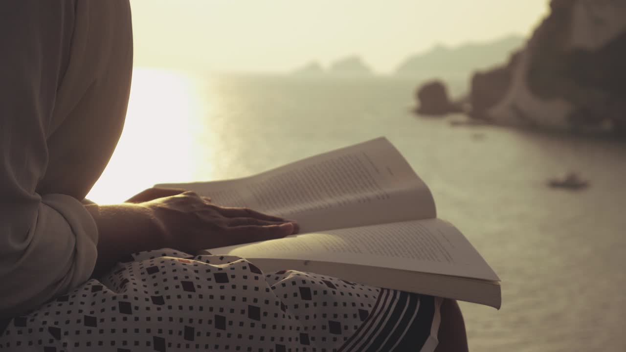 Young woman reading a book sitting on a wall at the beach at sunset on Ponza Island, Italy. Fashion white shirt and skirt.