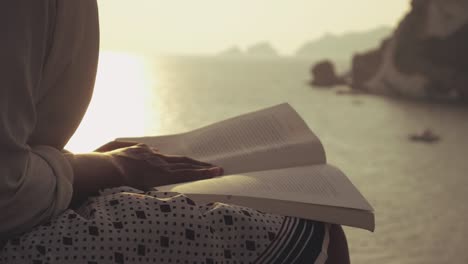 young woman reading a book sitting on a wall at the beach at sunset on ponza island, italy. fashion white shirt and skirt.