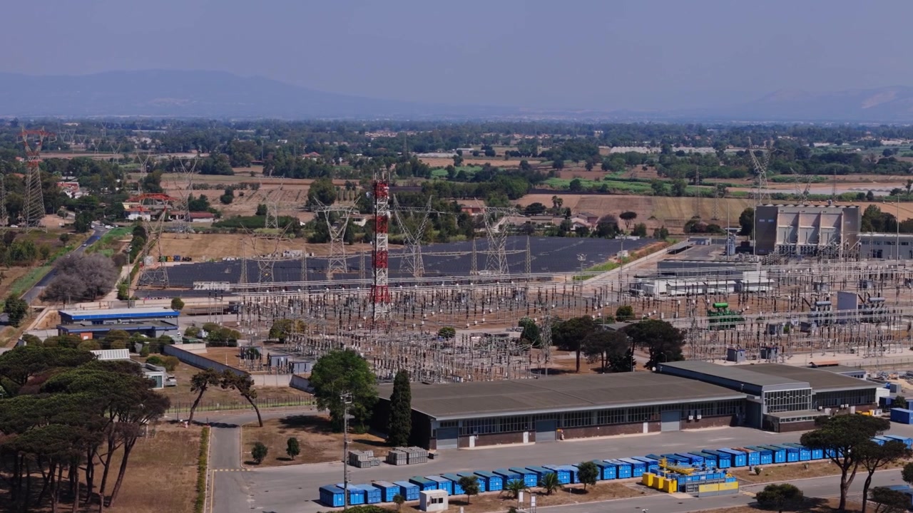 Vista de cima de uma grande subestação elétrica com linhas de alta tensão e torres ao lado de um parque solar gerando energia renovável.
