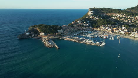 Puerto-Deportivo-Español-Soller-Al-Atardecer-Con-Barcos,-Océano-Azul-Y-Exuberantes-Montañas-Verdes-Toma-Panorámica-Aérea